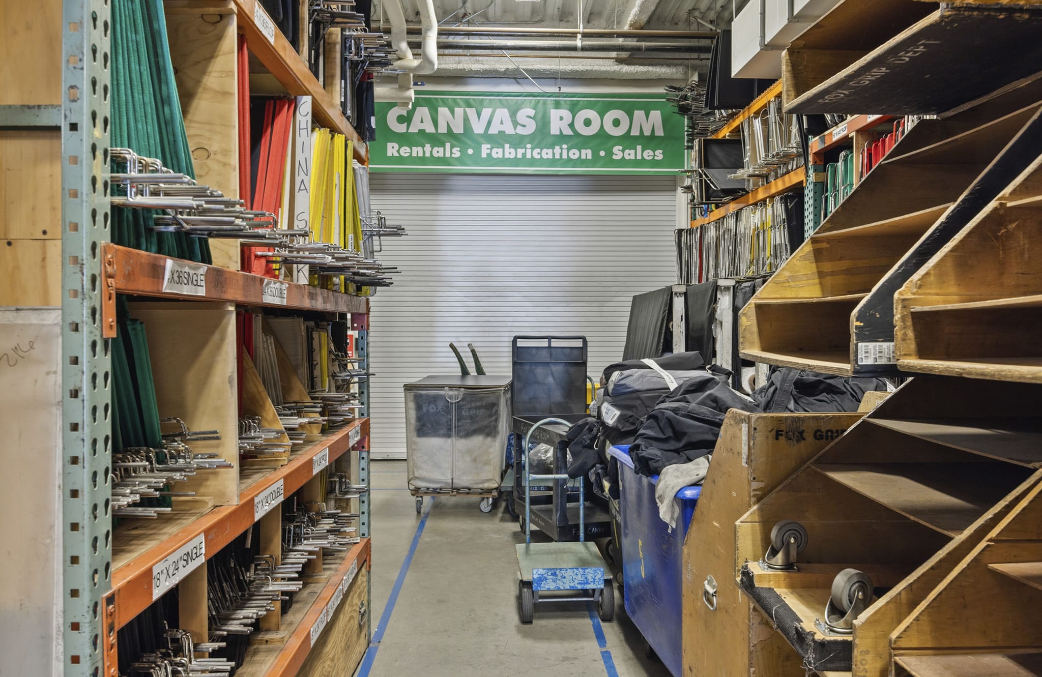 An aisle in a warehouse lined with shelving units holding various equipment and supplies, with a green "CANVAS ROOM" sign above a closed bay door.