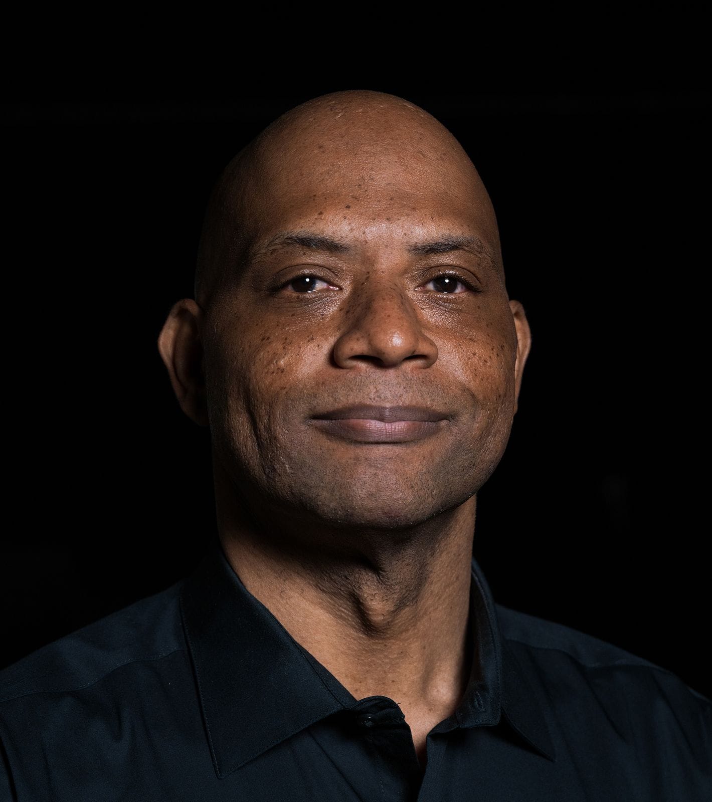 Headshot of a bald, dark-skinned man in a dark shirt, smiling gently at the camera.