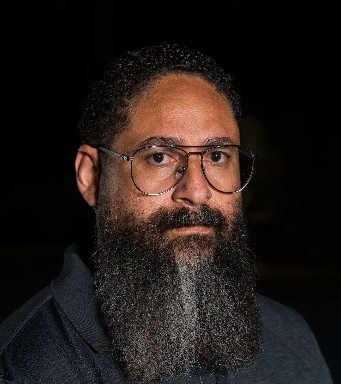 A man with dark curly hair, glasses, and a long grey-streaked beard looks directly at the camera.