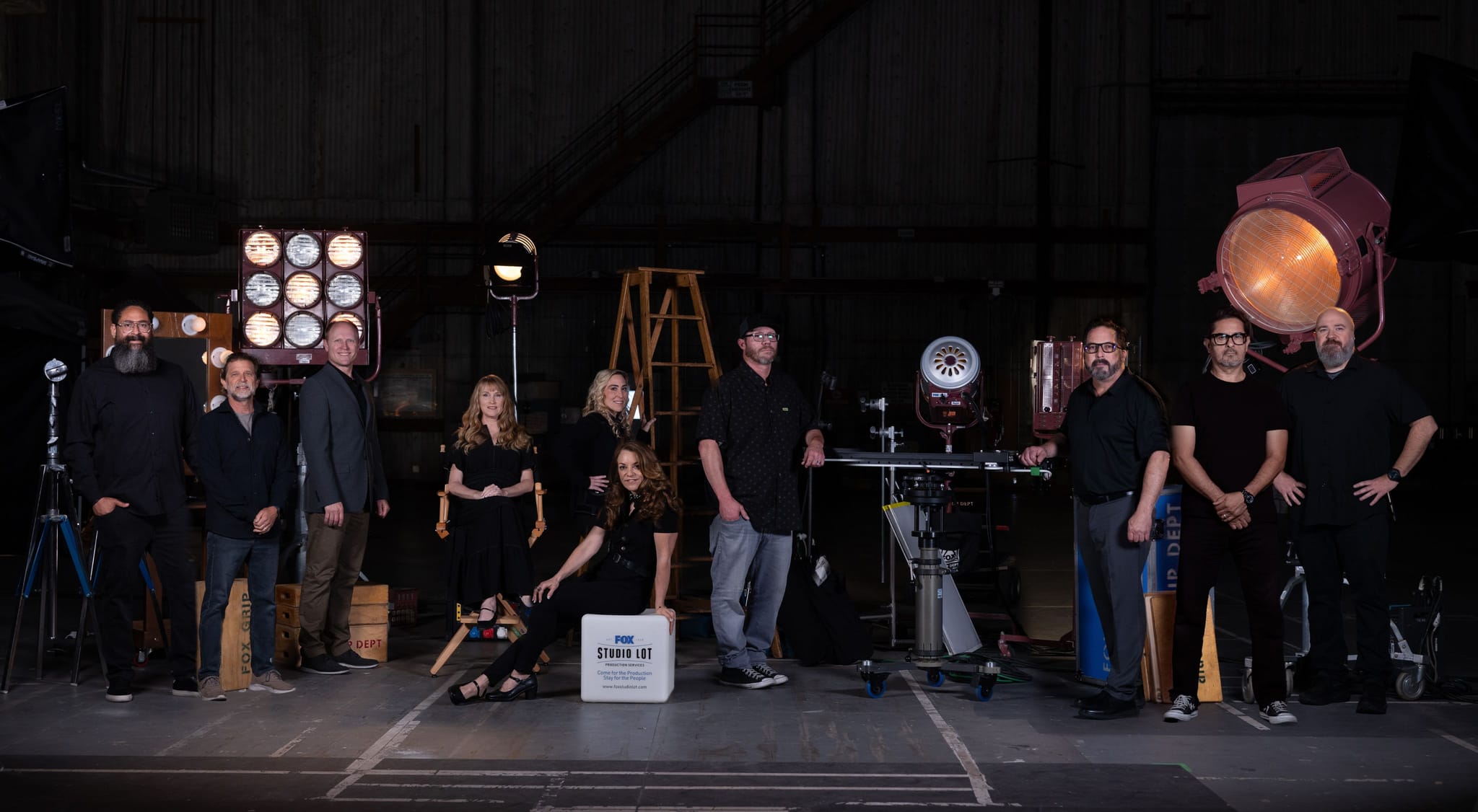 Ten film crew members pose among large studio lights and camera equipment in a dark soundstage.