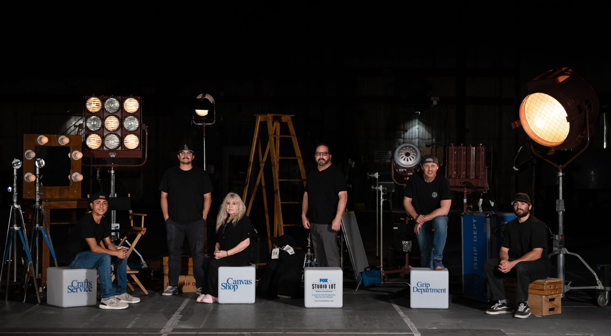Six film crew members pose on a dark set surrounded by lights and equipment, next to boxes labeled Crafts Service, Canvas Shop, PRO Studio L.I.T., and Grip Department.