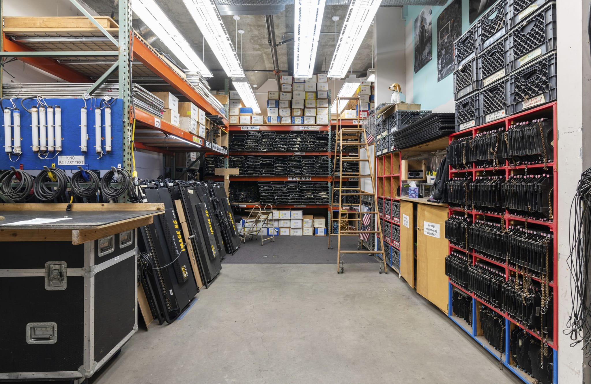 An organized warehouse filled with racks of stage equipment, including cables, cases, and stacked crates.