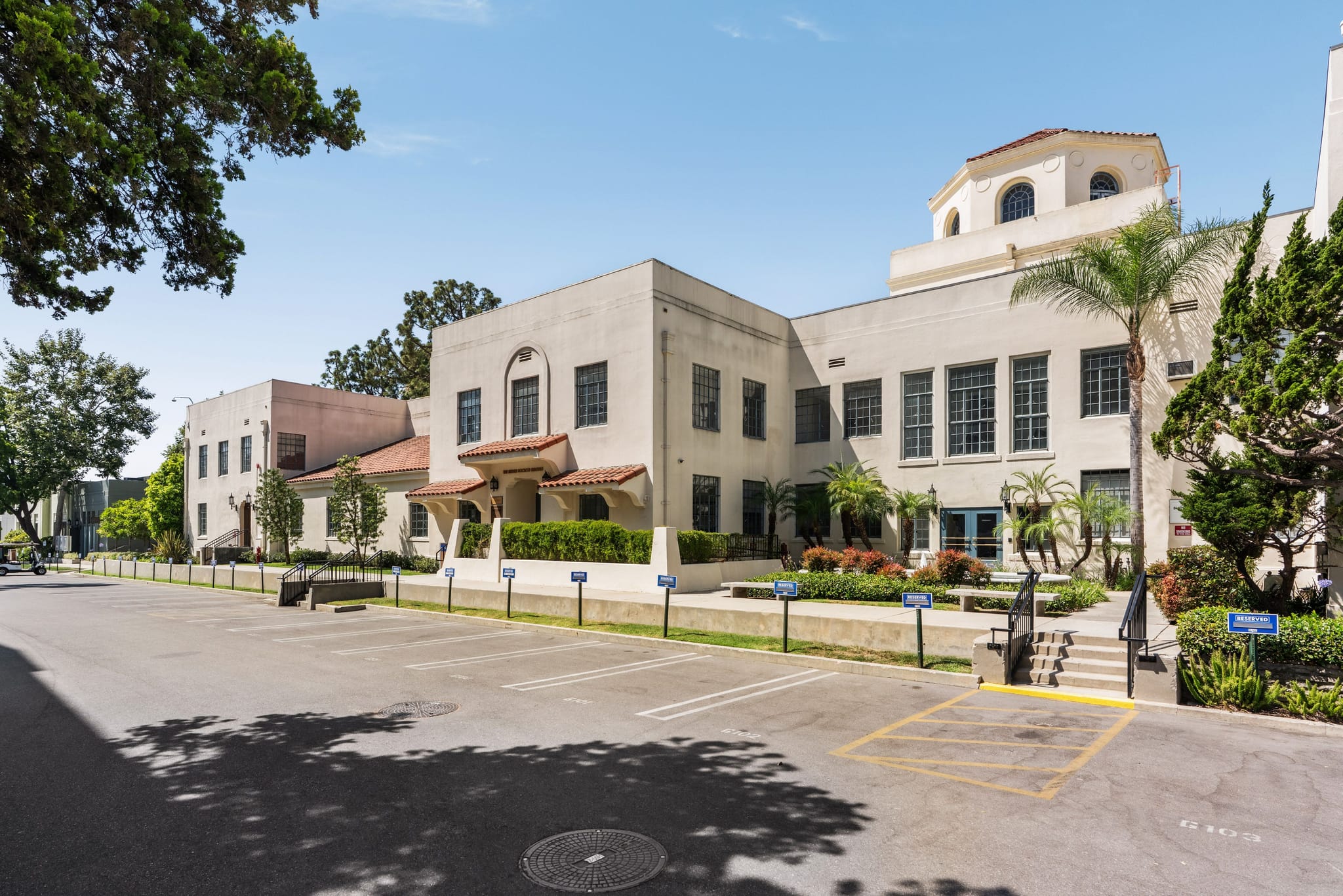 A large light-colored building with Spanish Colonial Revival architecture, a dome, and palm trees, fronted by an empty parking lot.