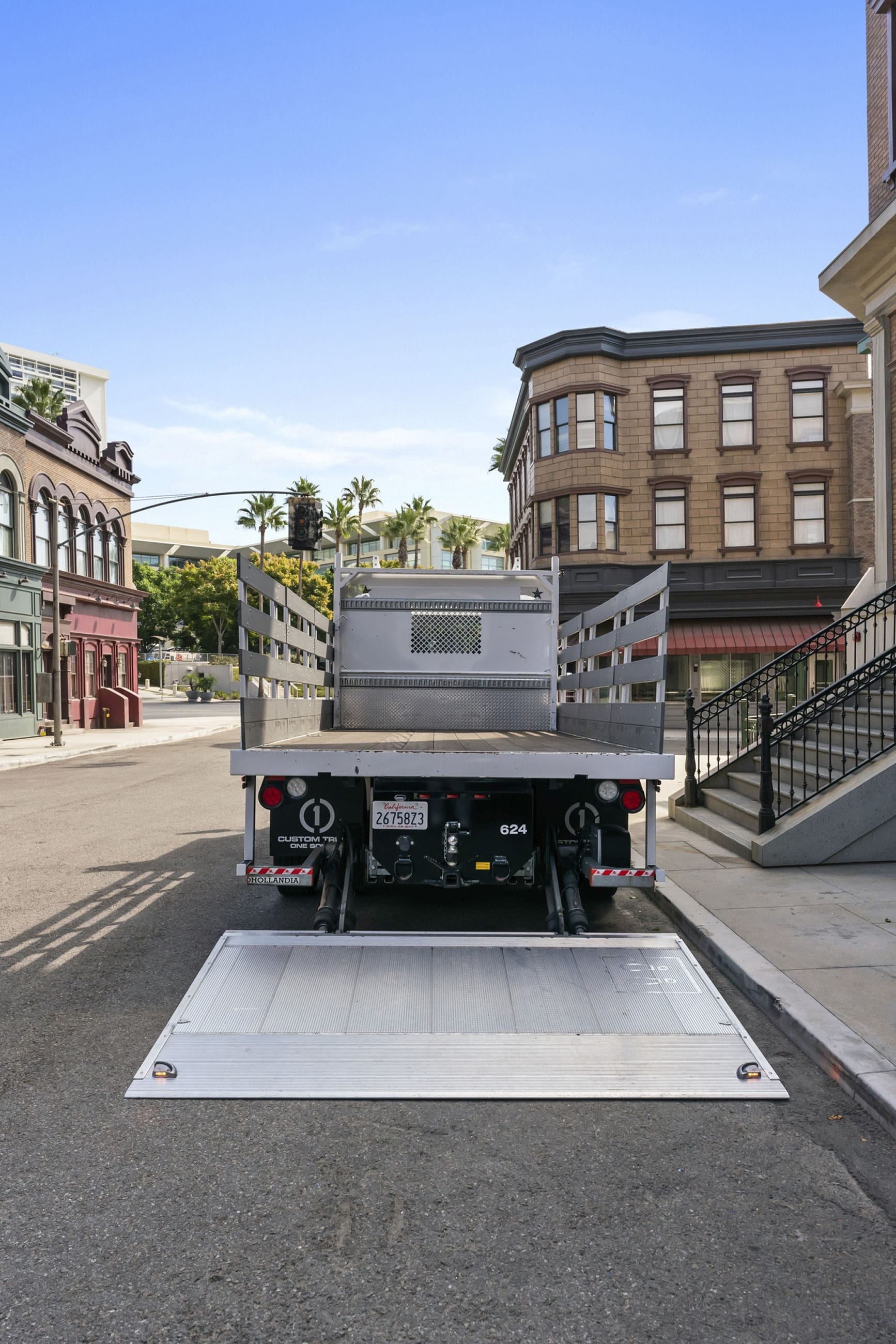 Rear view of a flatbed truck with its tailgate ramp lowered on a city street lined with buildings and palm trees.