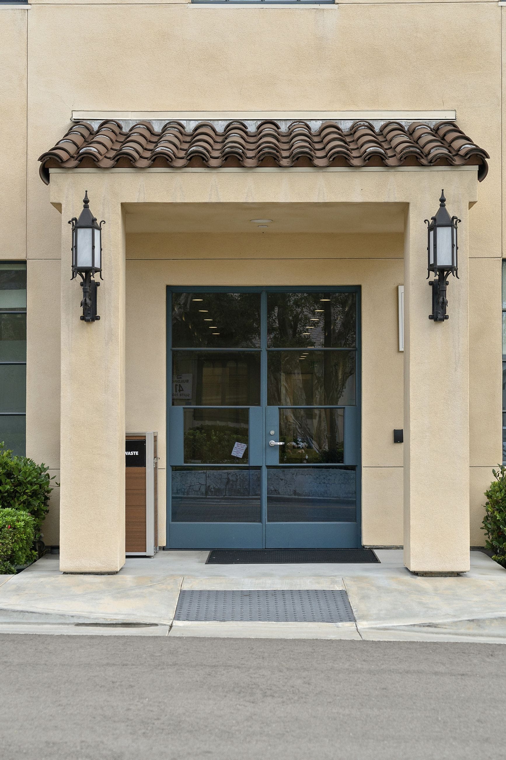 A building entrance with a beige facade, a brown tiled awning, and dark teal double doors flanked by black wall lanterns.