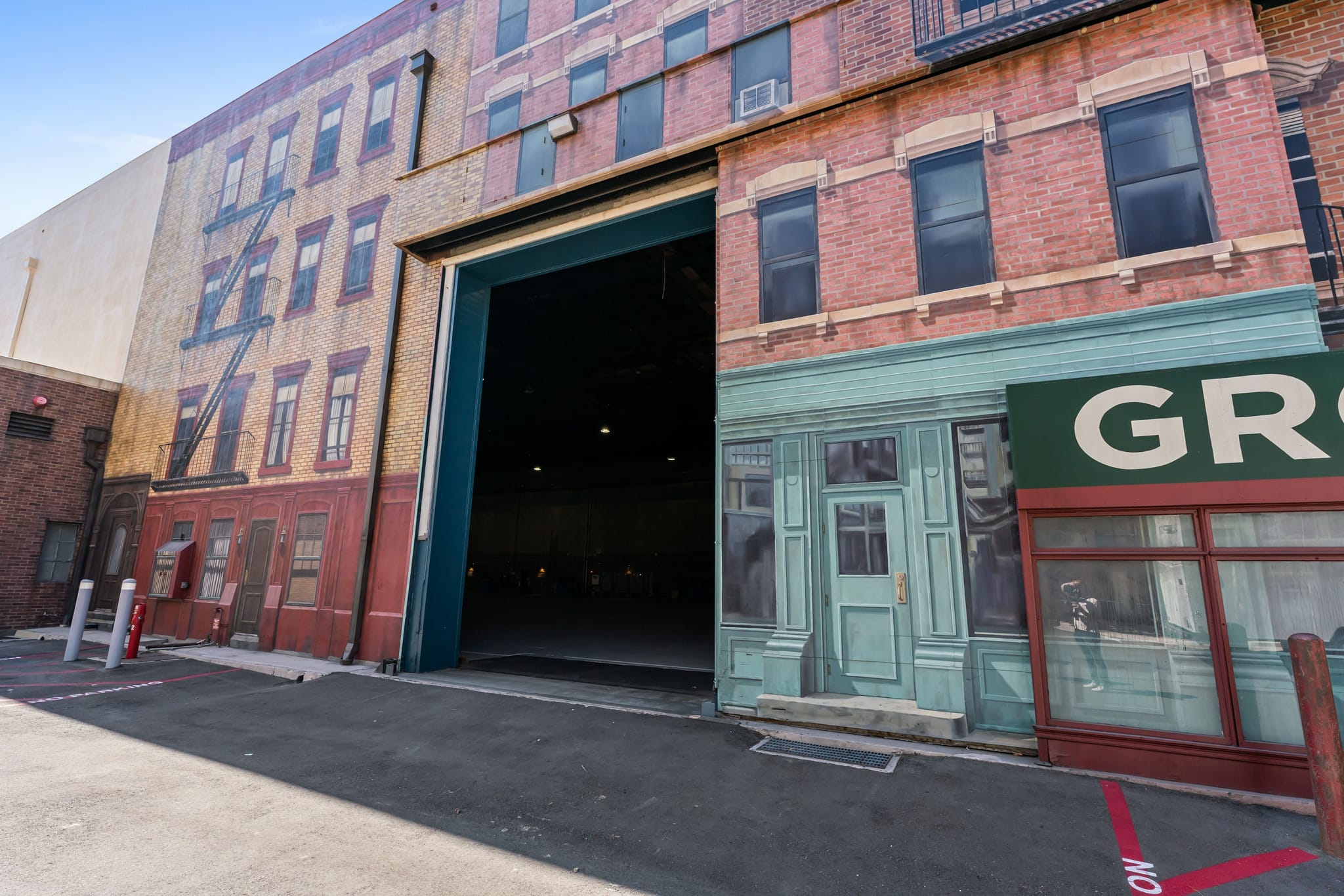 Studio facade designed as a city street, featuring painted brick buildings, a large open soundstage entrance, and storefronts including a partial 'GROCERY' sign.