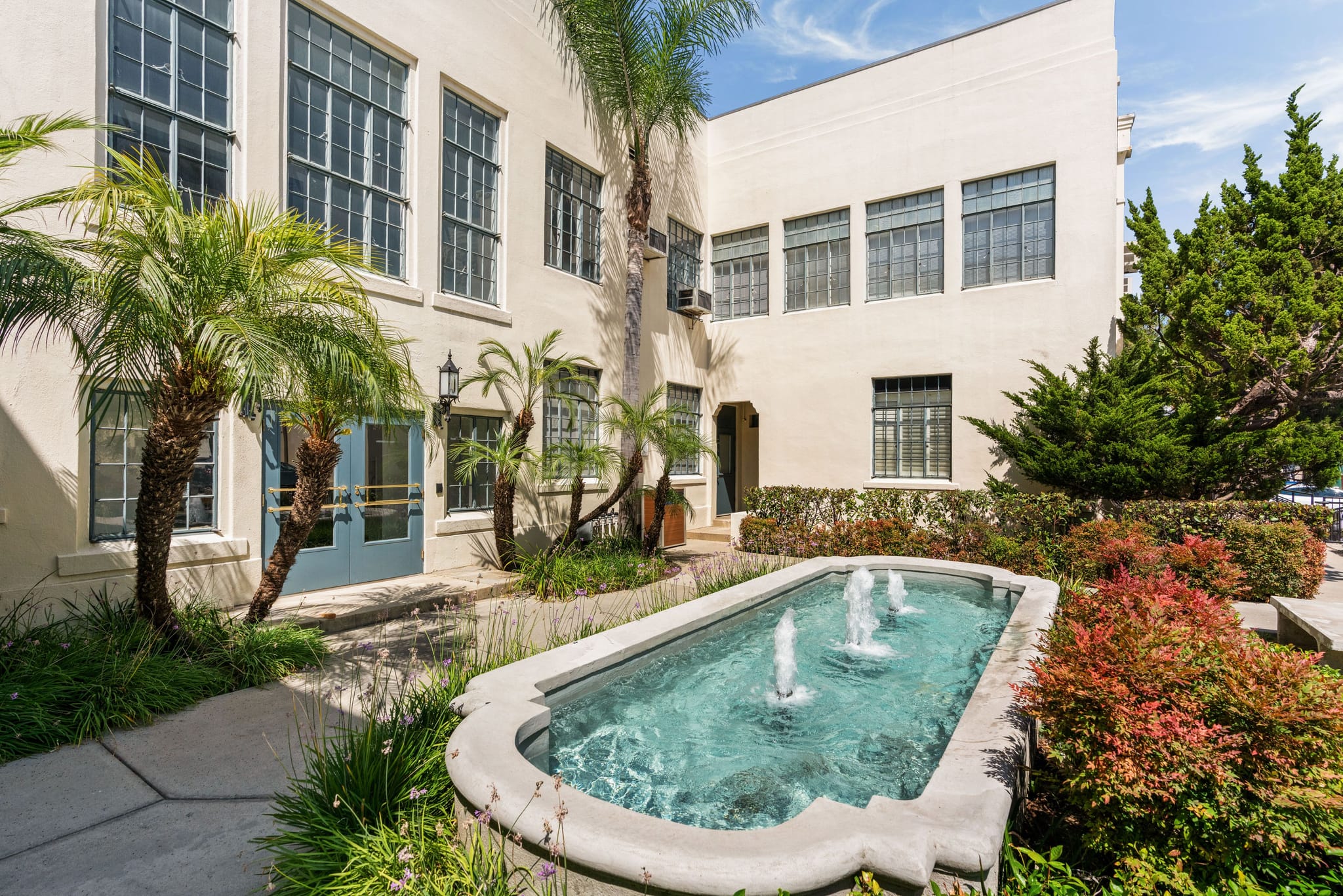 An outdoor courtyard featuring a rectangular fountain with three jets, surrounded by palm trees and colorful shrubs, in front of a light-colored building with many windows.