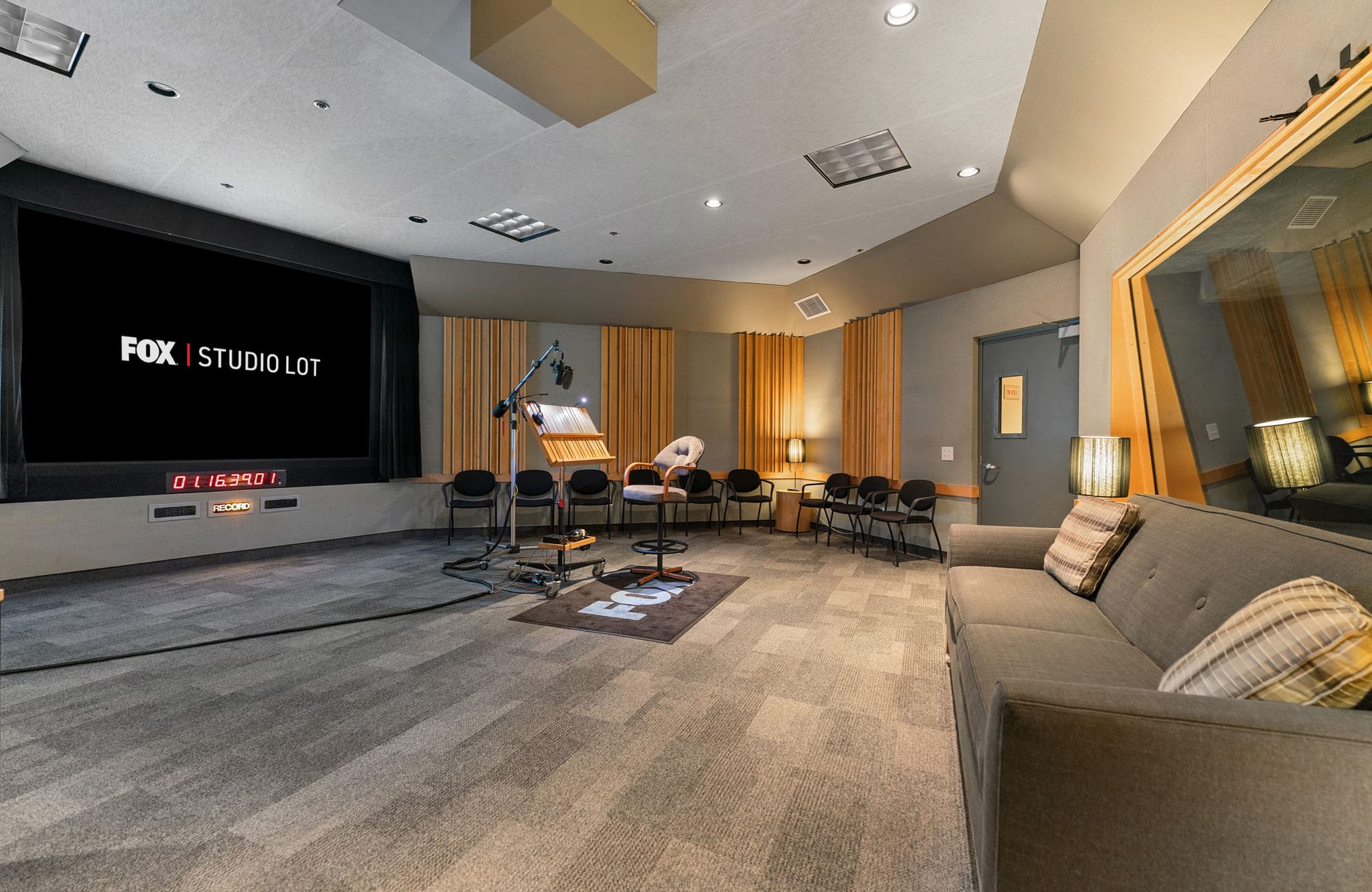 A professional audio recording studio with a large screen displaying 'FOX | STUDIO LOT', microphones, and seating.