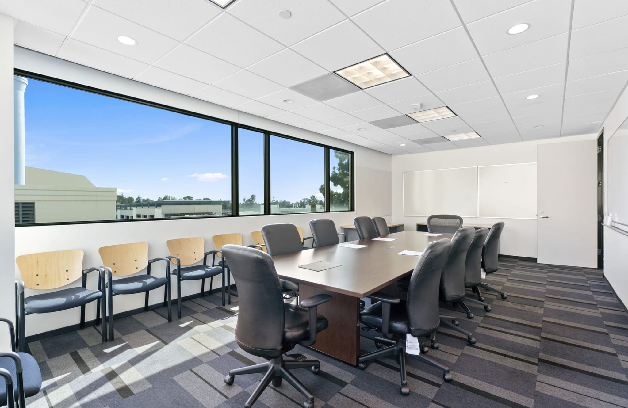 A modern conference room with a long table, office chairs, a large window overlooking an outdoor scene, and whiteboards.