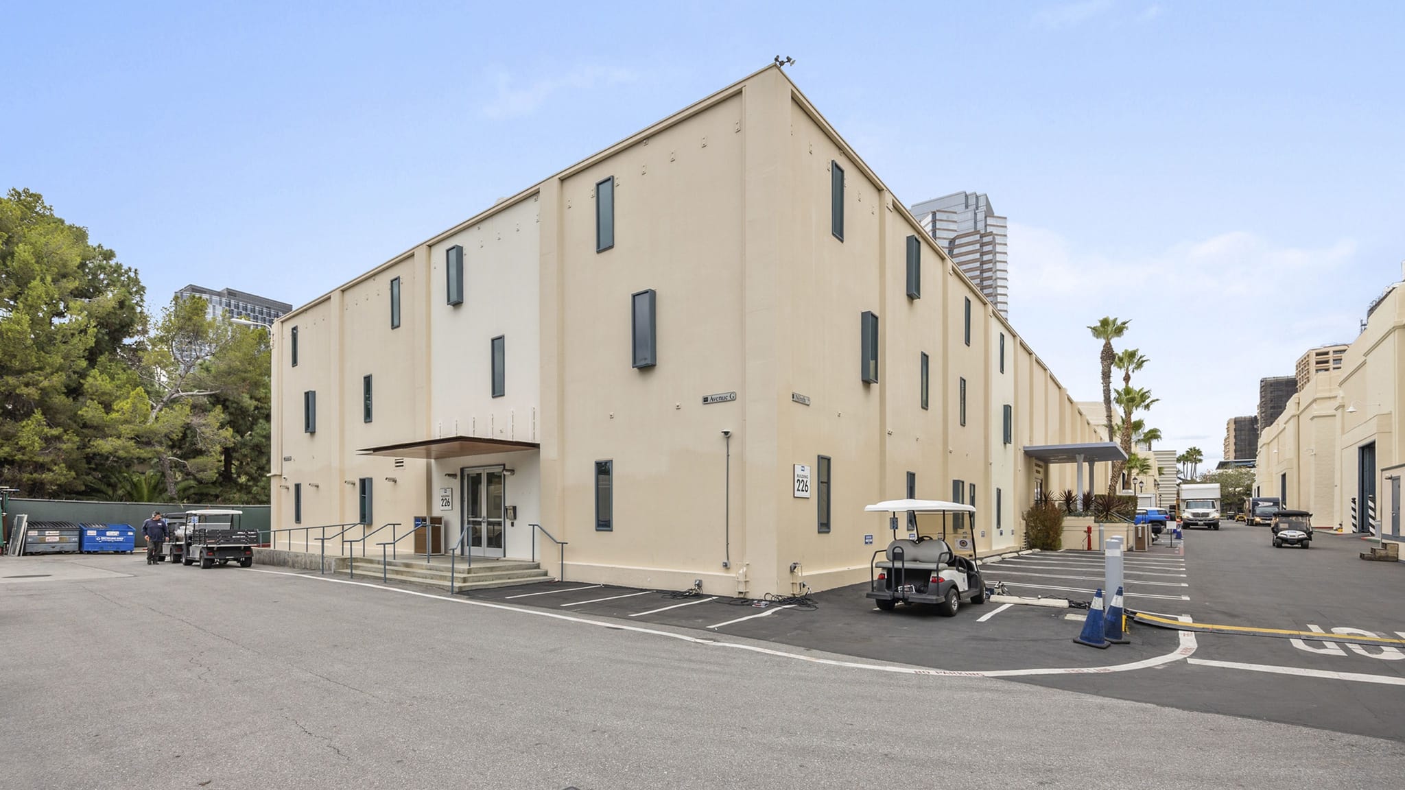 A large, light-colored two-story studio building with tall windows, a paved lot with a golf cart, and other buildings in the background.