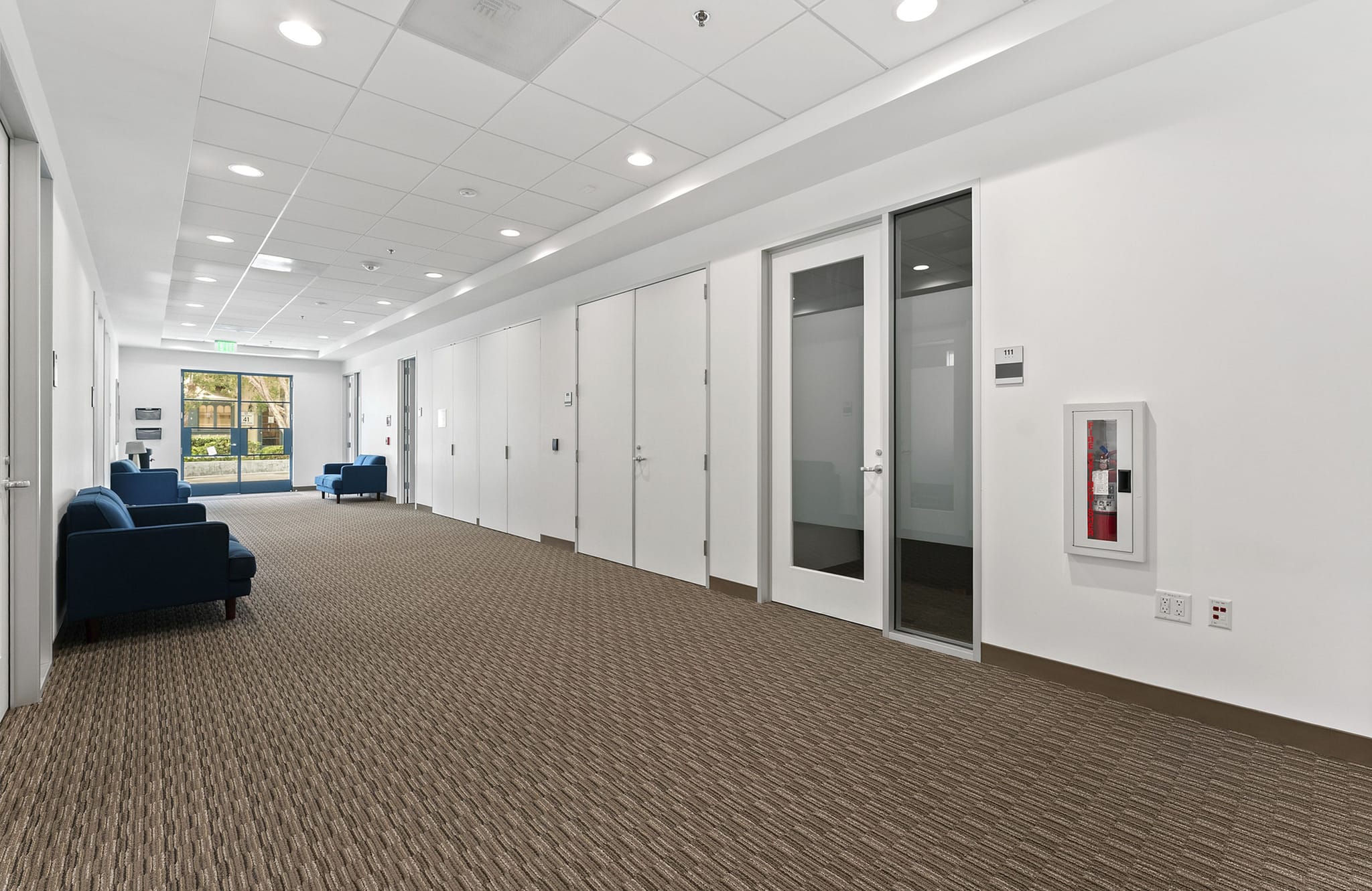 Bright, clean hallway with white walls, brown patterned carpet, blue seating, multiple doors, and an exterior view at the end.