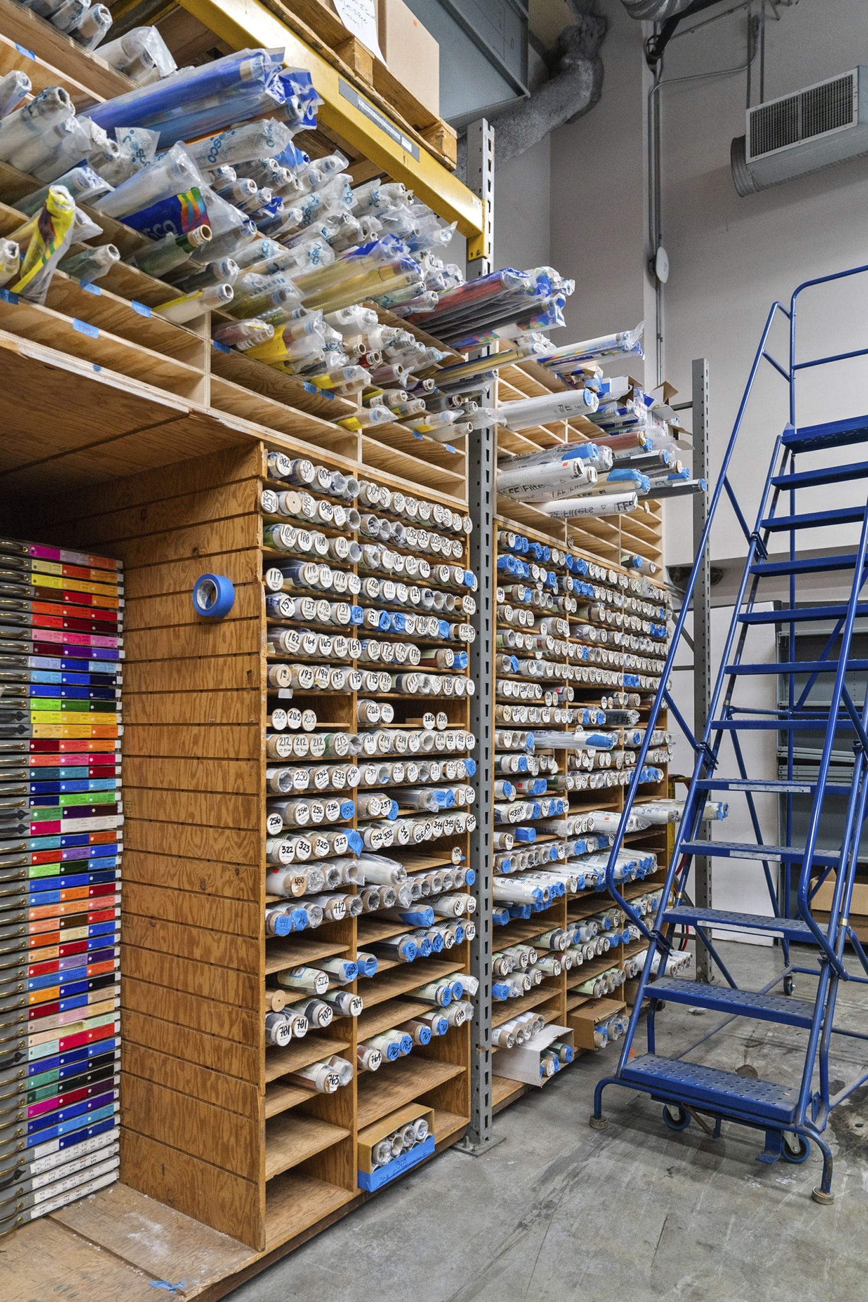 Extensive storage racks filled with rolls of wrapped materials, beside a blue rolling ladder.