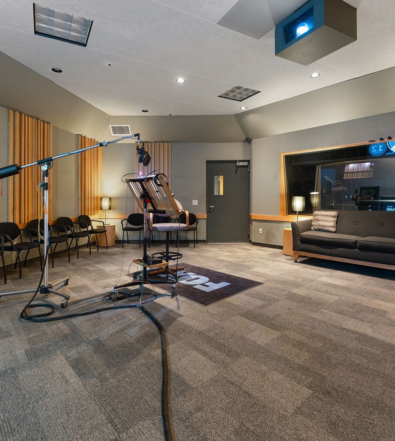 A professional recording studio with a microphone stand, script stand, a dark sofa, and a window looking into a control room.