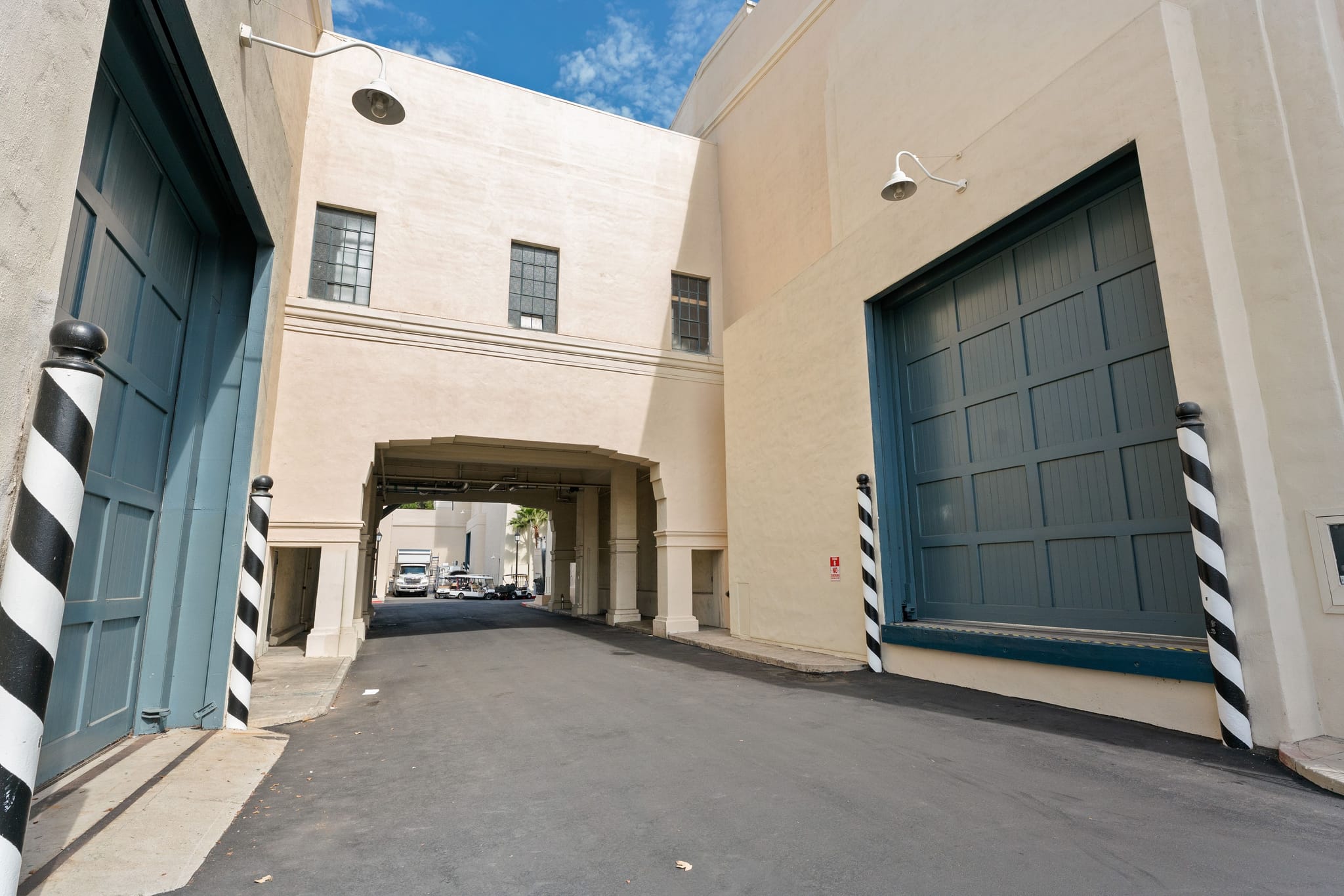 A paved passage between two light-colored buildings with large blue garage doors flanked by striped bollards, and an archway connecting the buildings overhead.