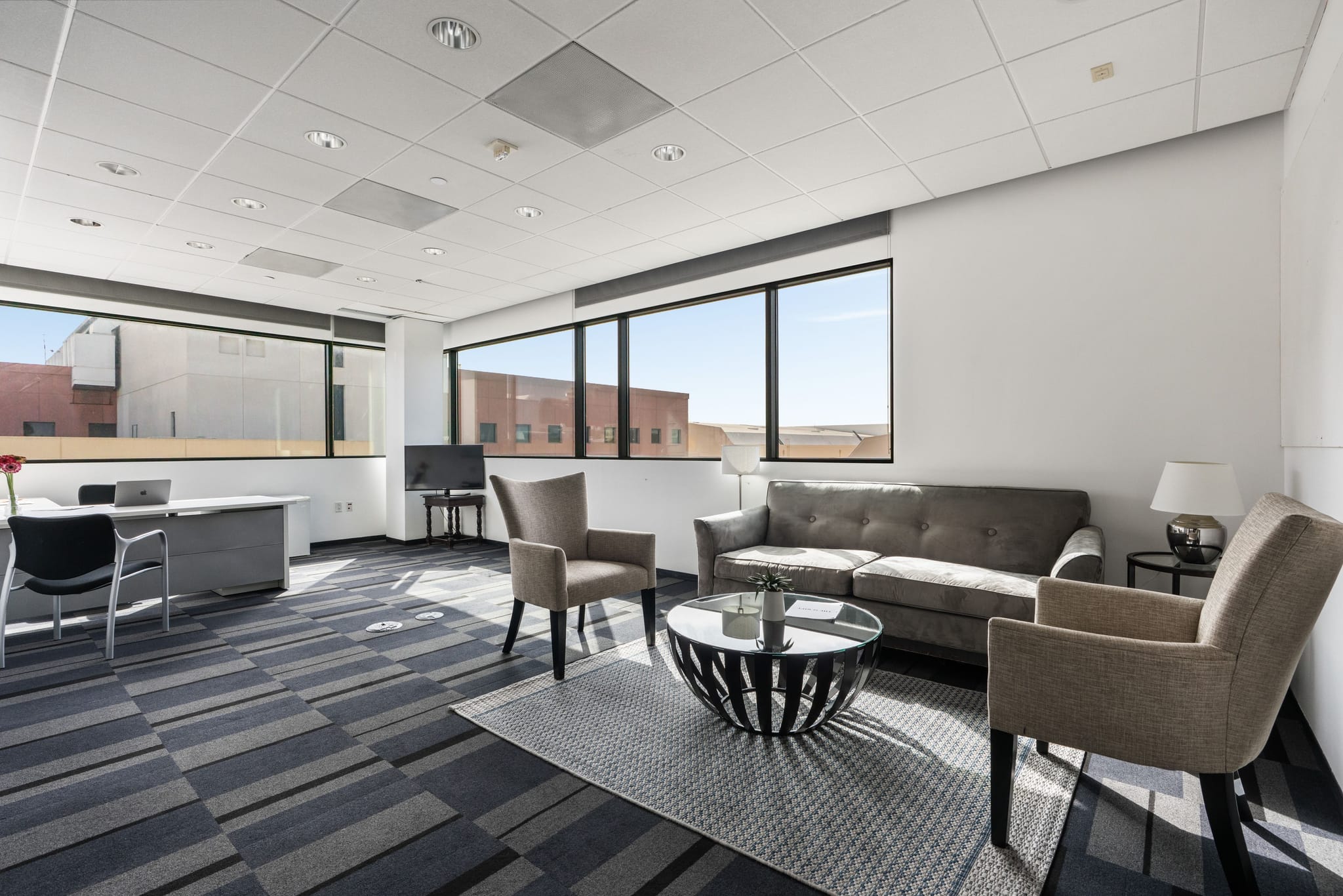 A modern office space with a gray sofa, two armchairs, a coffee table, a desk with a laptop, and large windows.