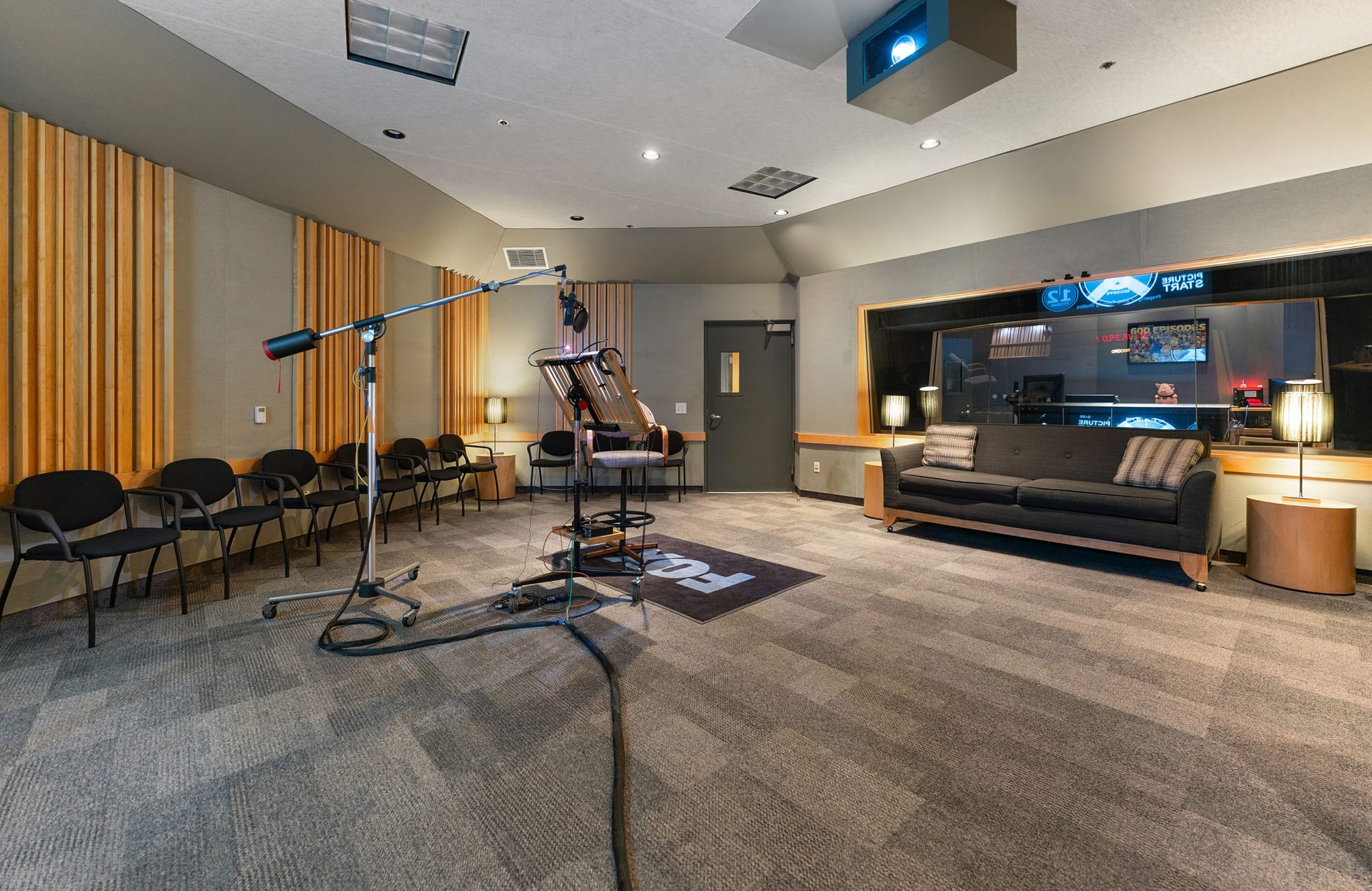 A professional recording studio with a microphone setup, a sofa, and a large window looking into a control room.
