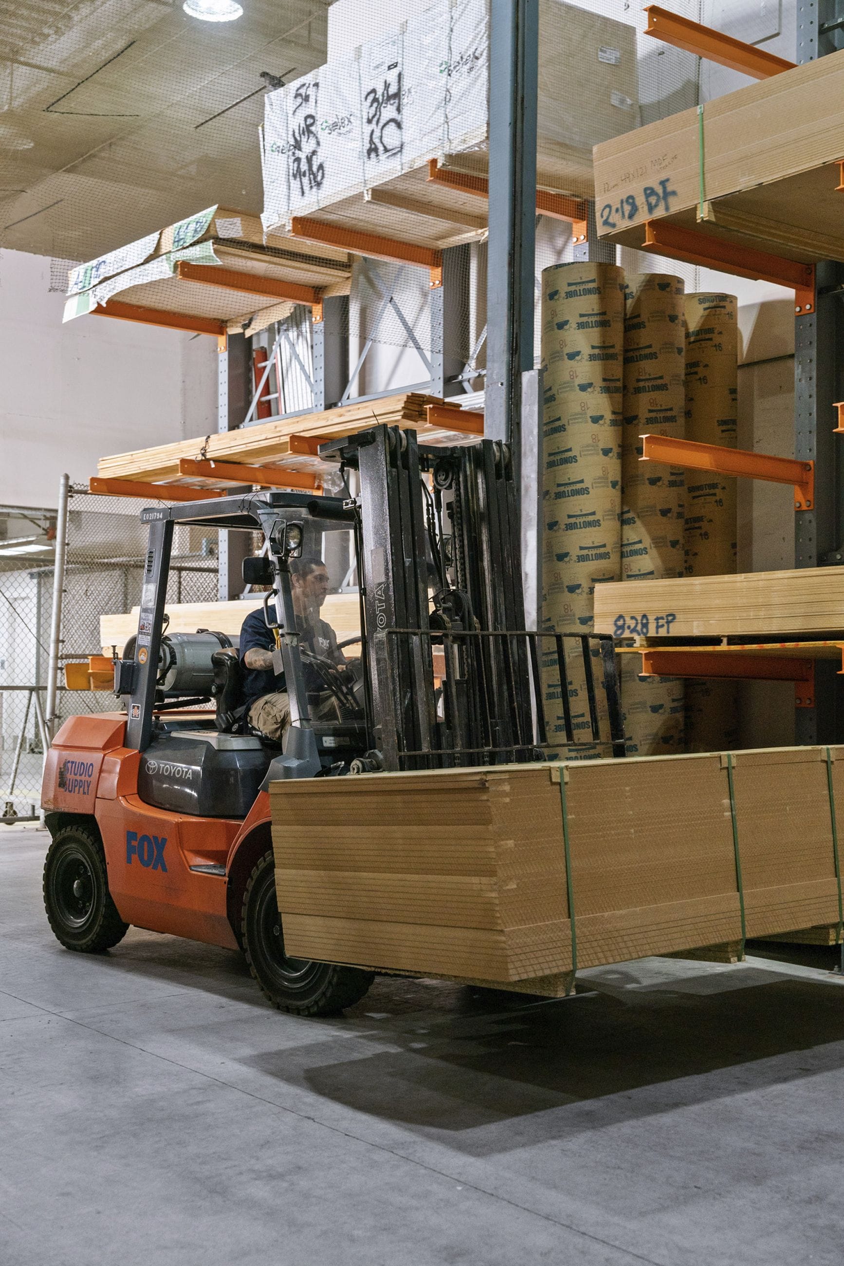 A person operates an orange forklift carrying a large stack of wood panels in a warehouse.