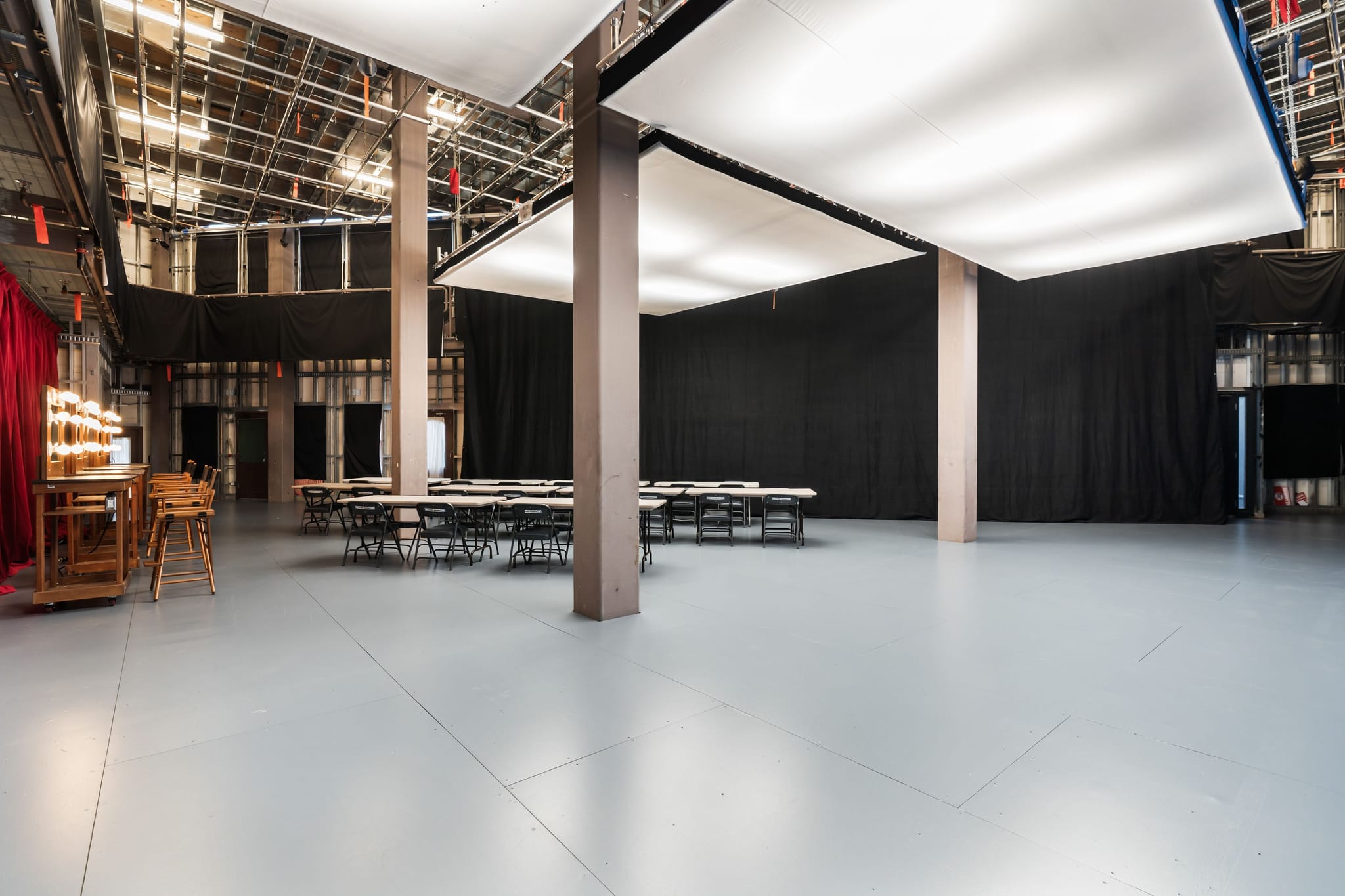 Large studio with black drapes, overhead lighting panels, a makeup vanity, and tables with chairs.