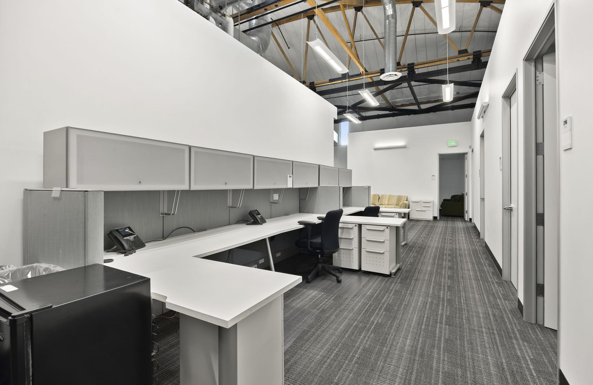 A modern office space with several workstations, an exposed industrial-style ceiling, and a hallway with multiple doors.