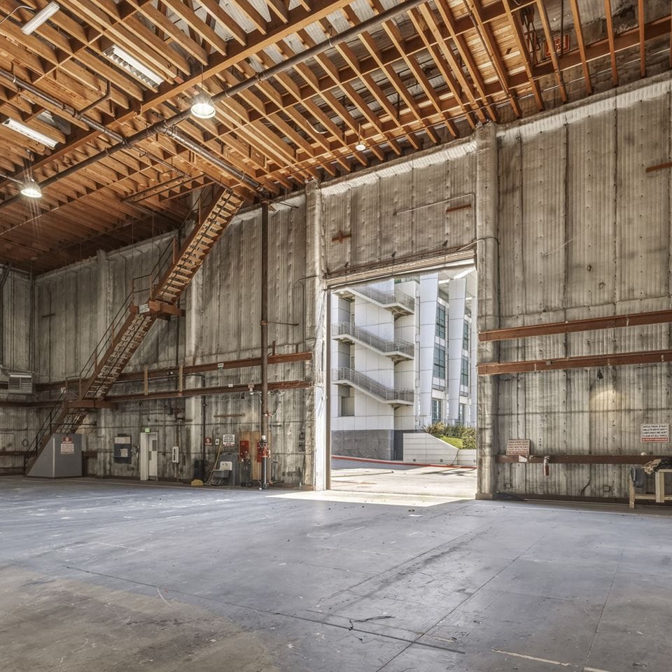 Empty industrial space with concrete walls, exposed wooden ceiling, and an open doorway revealing a modern building with stairs.