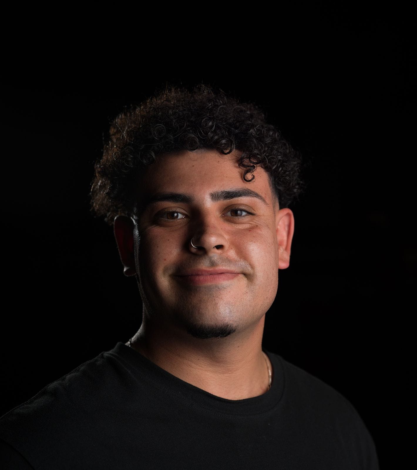 A young man with curly dark hair and a nose ring smiles at the camera.