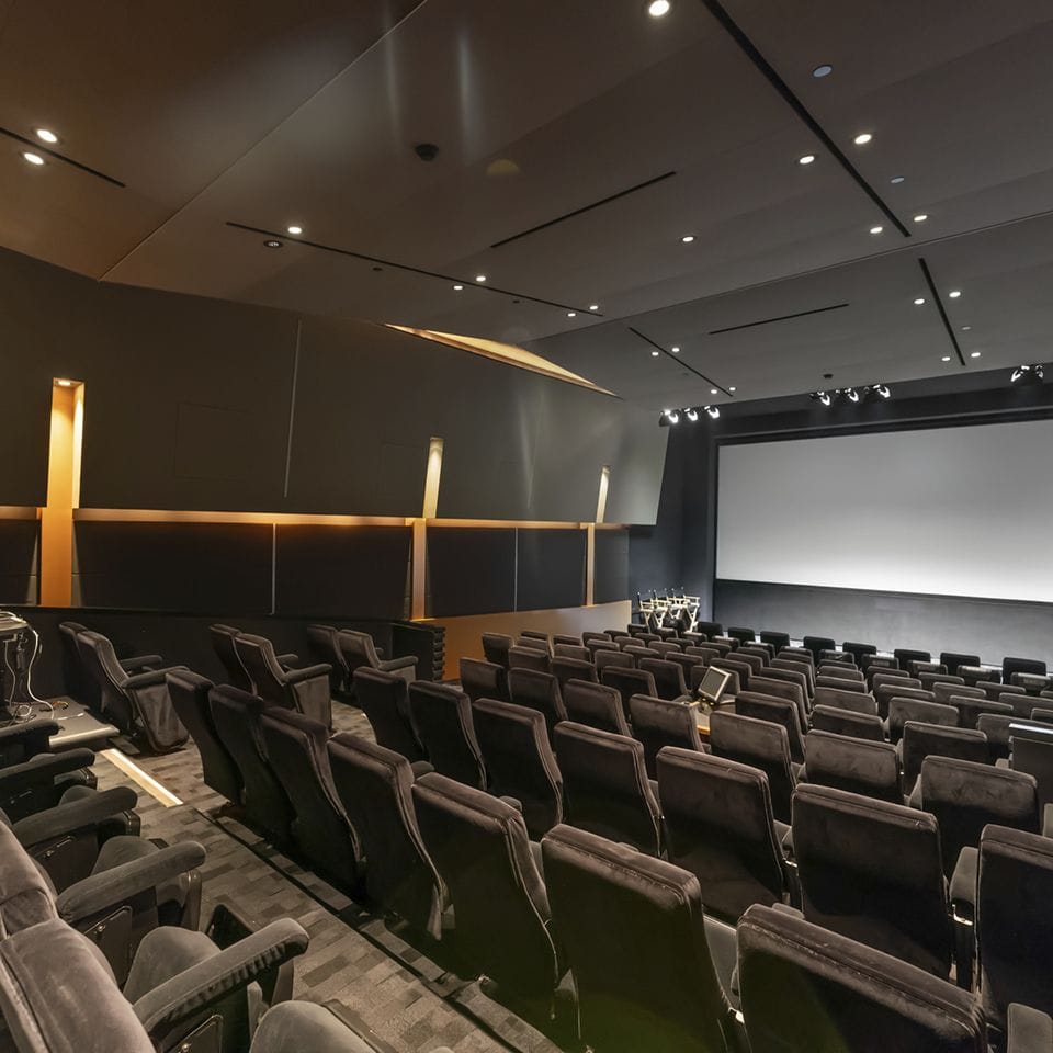 An empty, dark-themed modern auditorium with rows of plush seats facing a large projection screen.