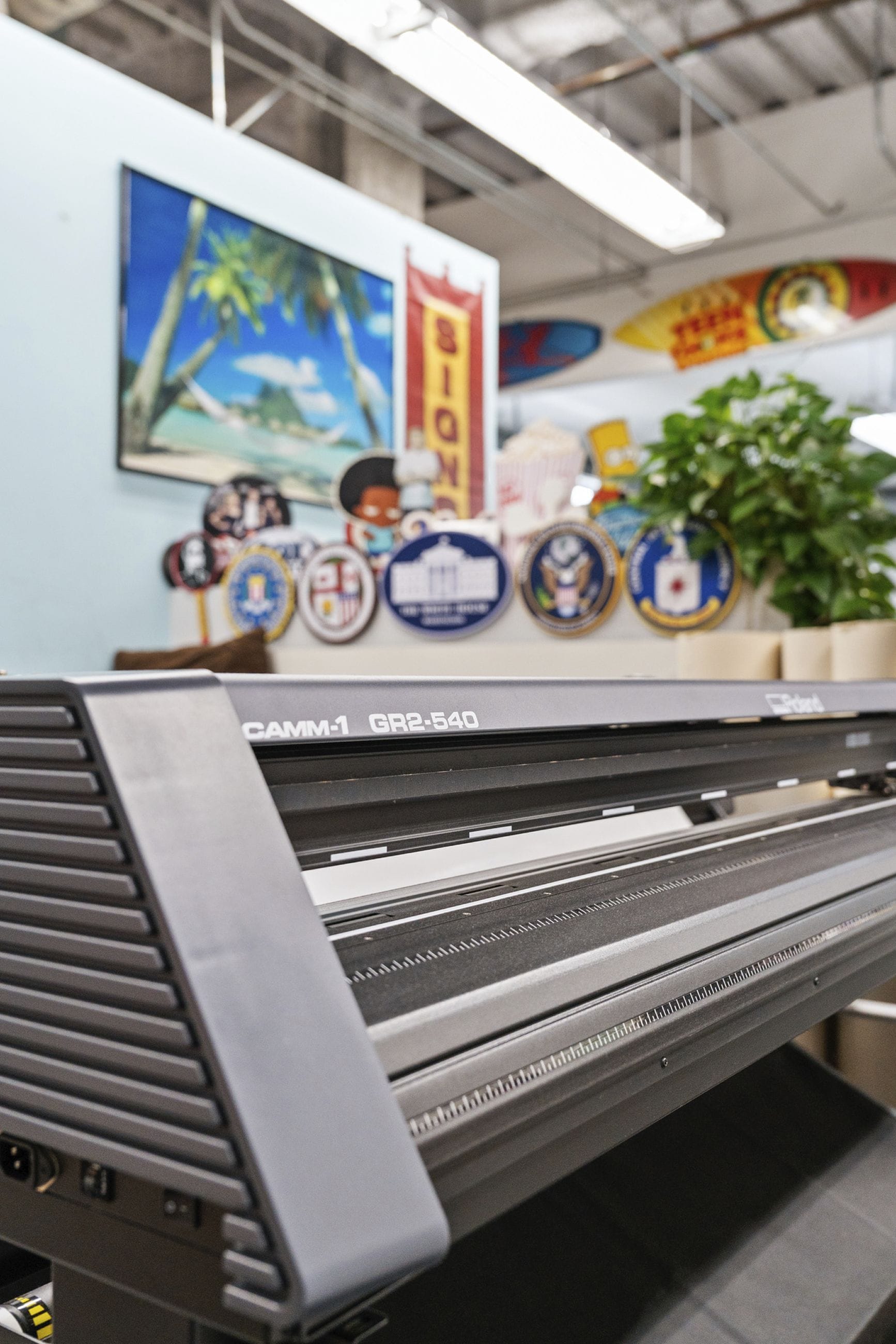 A Roland CAMM-1 GR2-540 vinyl cutter in the foreground, with blurry promotional badges, a beach poster, and a banner in the background.