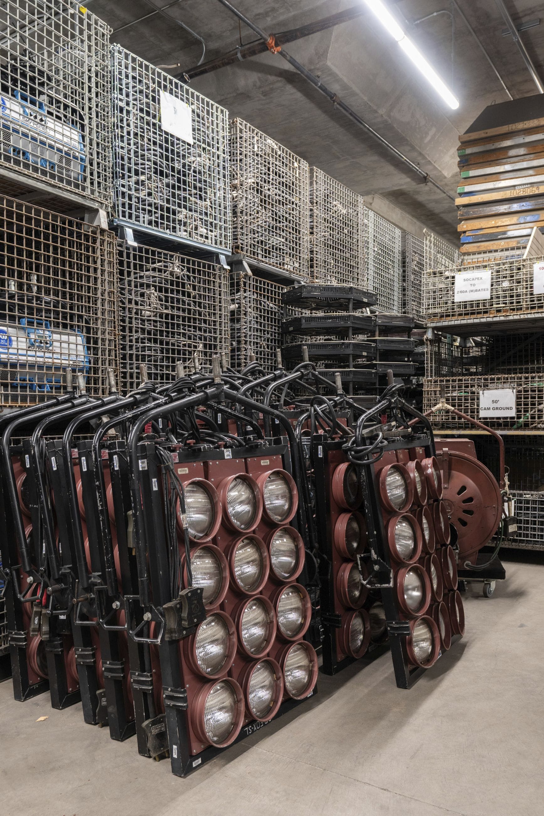Reddish-brown multi-bulb stage lights stand in a warehouse with stacked wire mesh crates of equipment behind them.