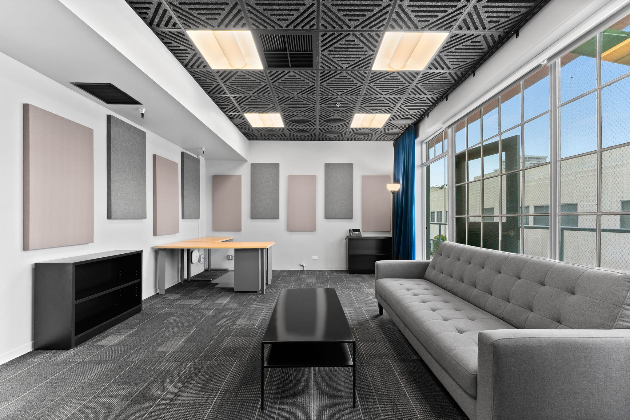 Modern studio or office lounge with acoustic wall panels, a patterned ceiling, a grey sofa, black coffee table, and large windows.