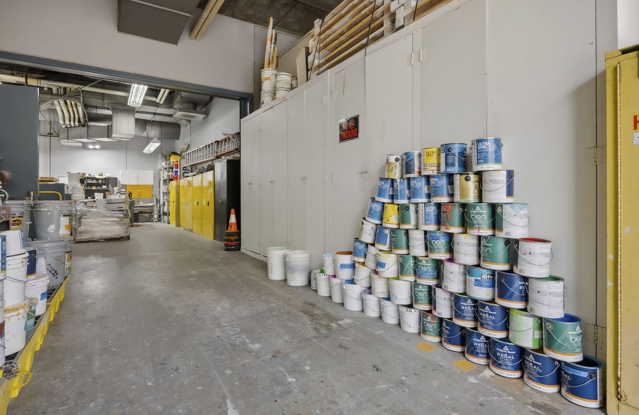 A storage room with a large pyramid stack of colorful paint cans on the right and an aisle leading to supplies and equipment on the left.