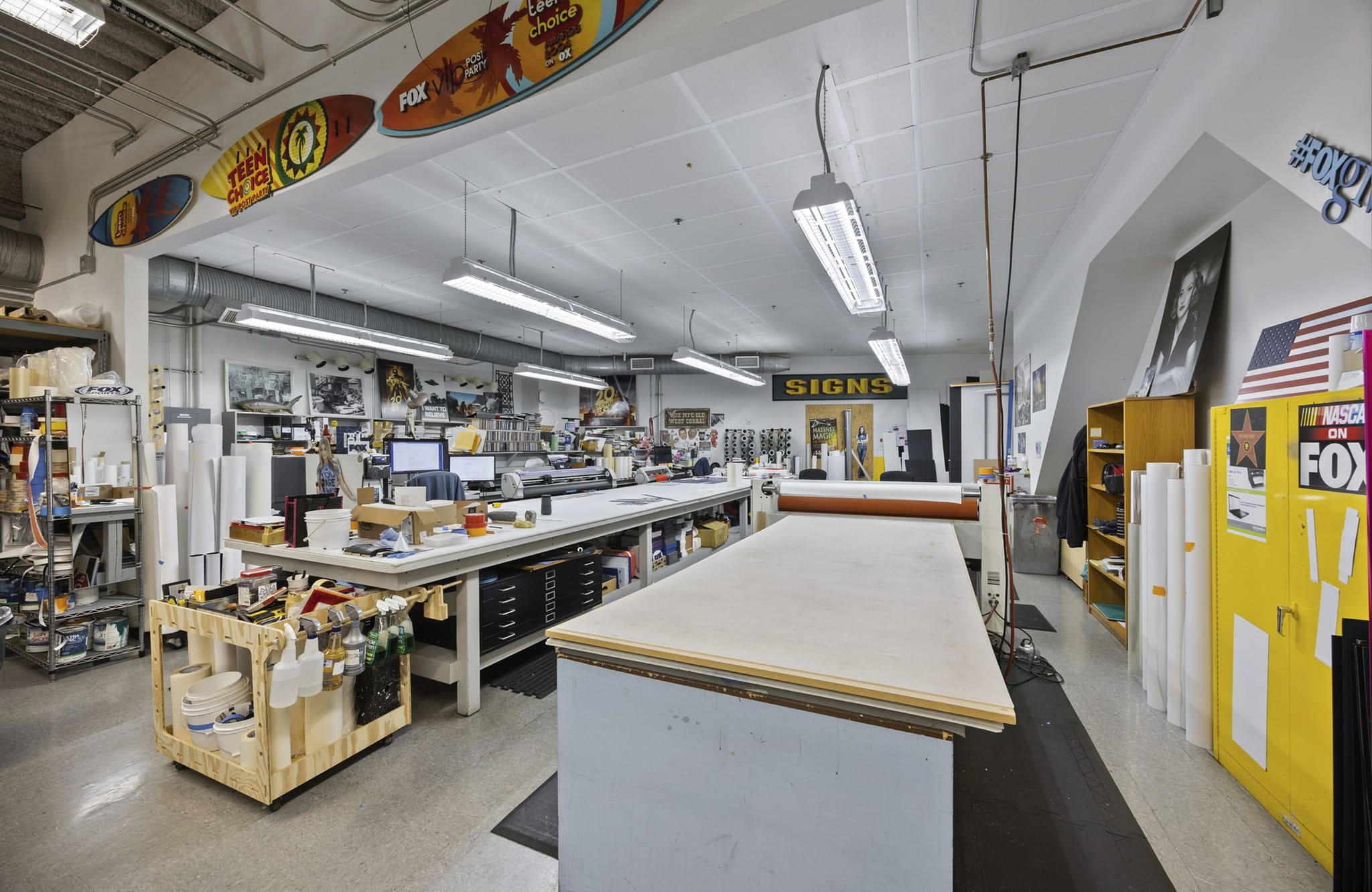 Interior of a busy sign and print shop with work tables, equipment, and rolls of material.