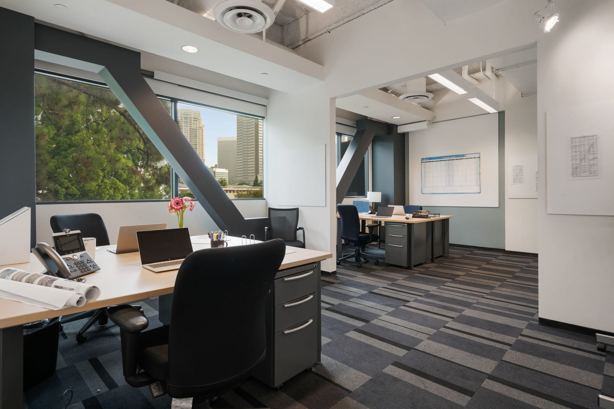 A modern, open-plan office space with multiple desks, chairs, computers, and large windows overlooking trees and city buildings.