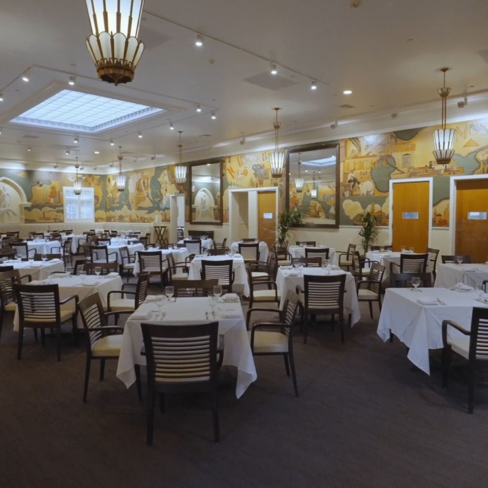 Empty, elegant dining room with white-clothed tables, colorful wall murals, and ornate chandeliers.