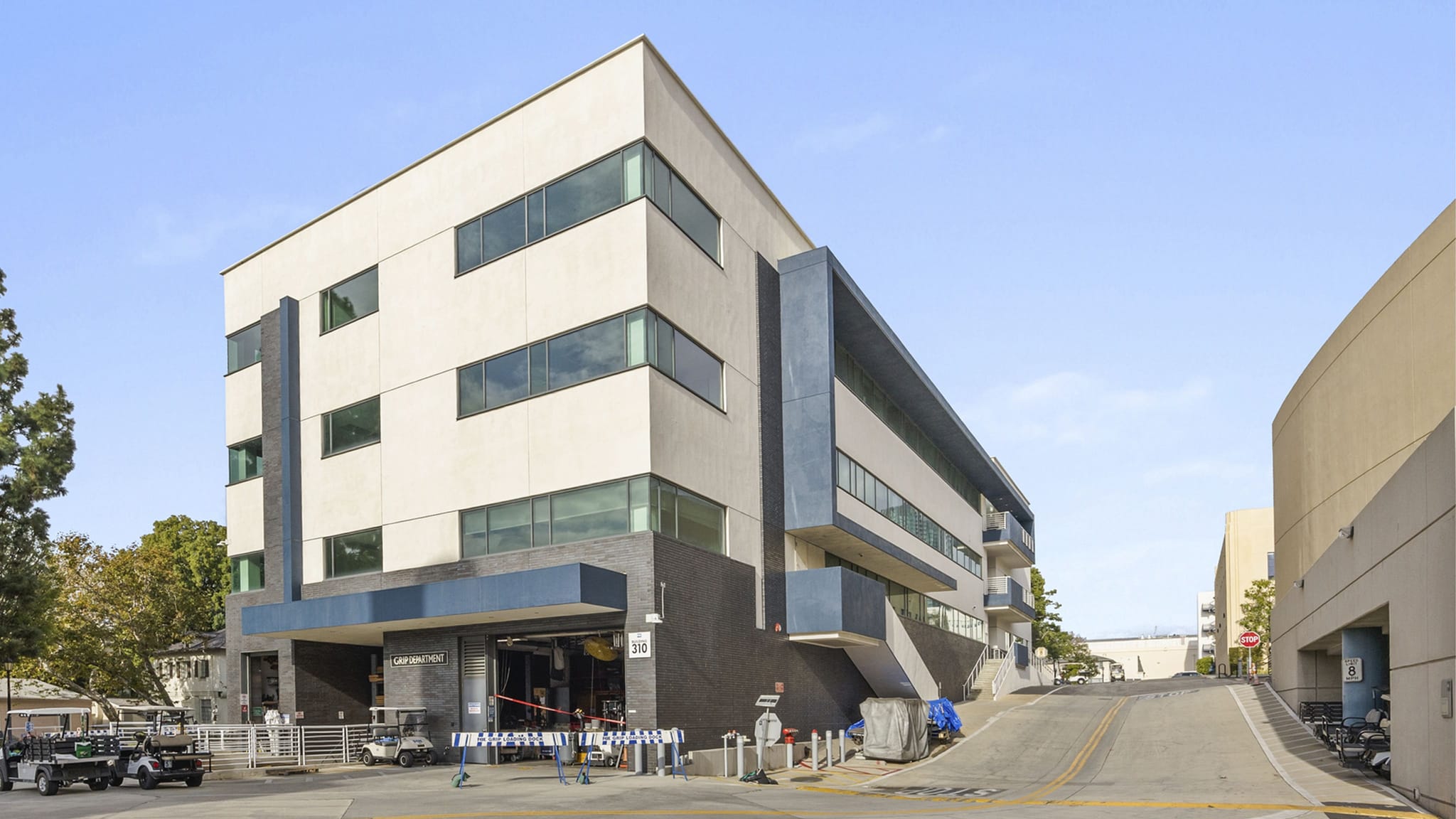 A modern, multi-story building with light-colored walls and dark blue accents, flanked by an uphill paved road.