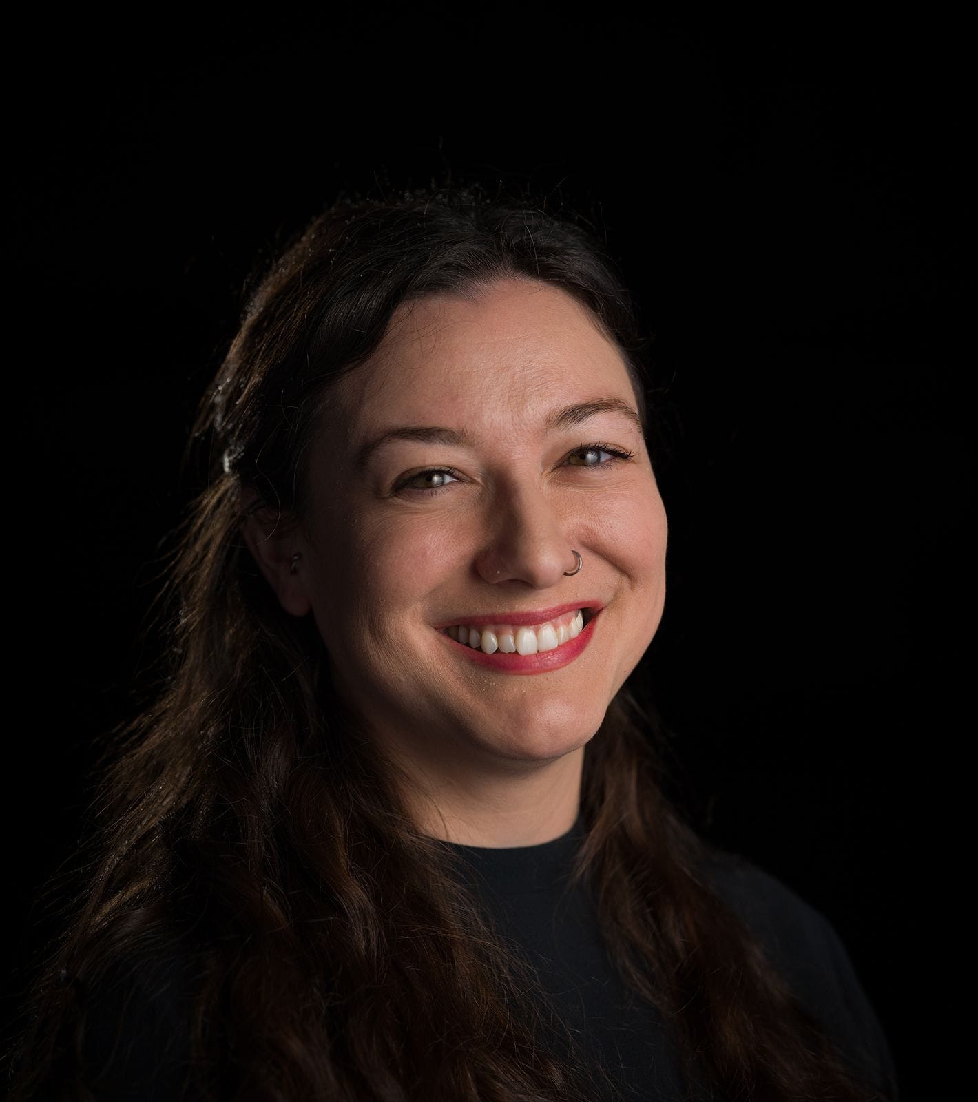 A smiling woman with long dark hair, red lipstick, and a nose ring against a black background.