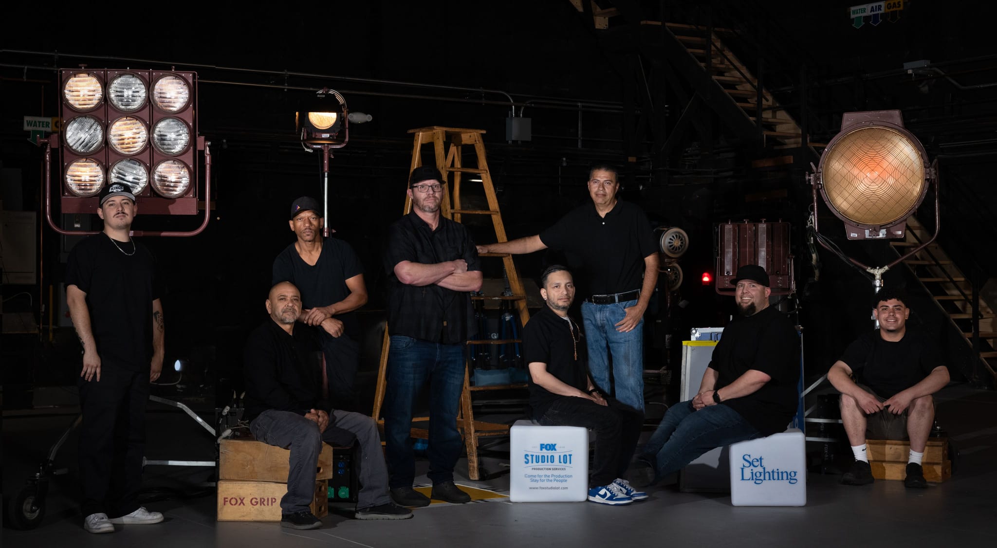 Eight men pose in a dark studio surrounded by large lights, ladders, and production boxes.