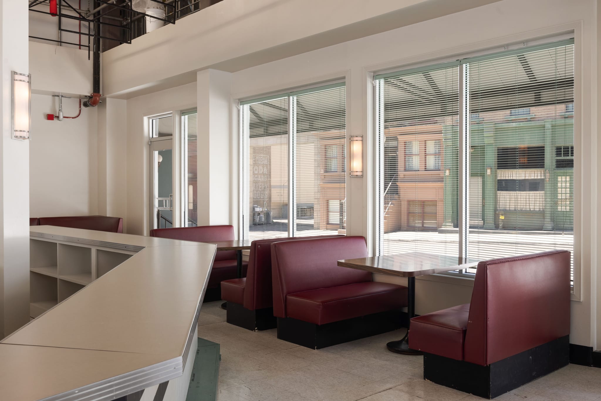 Interior of a retro diner with red booths, a long counter, and large windows looking out onto a street scene.