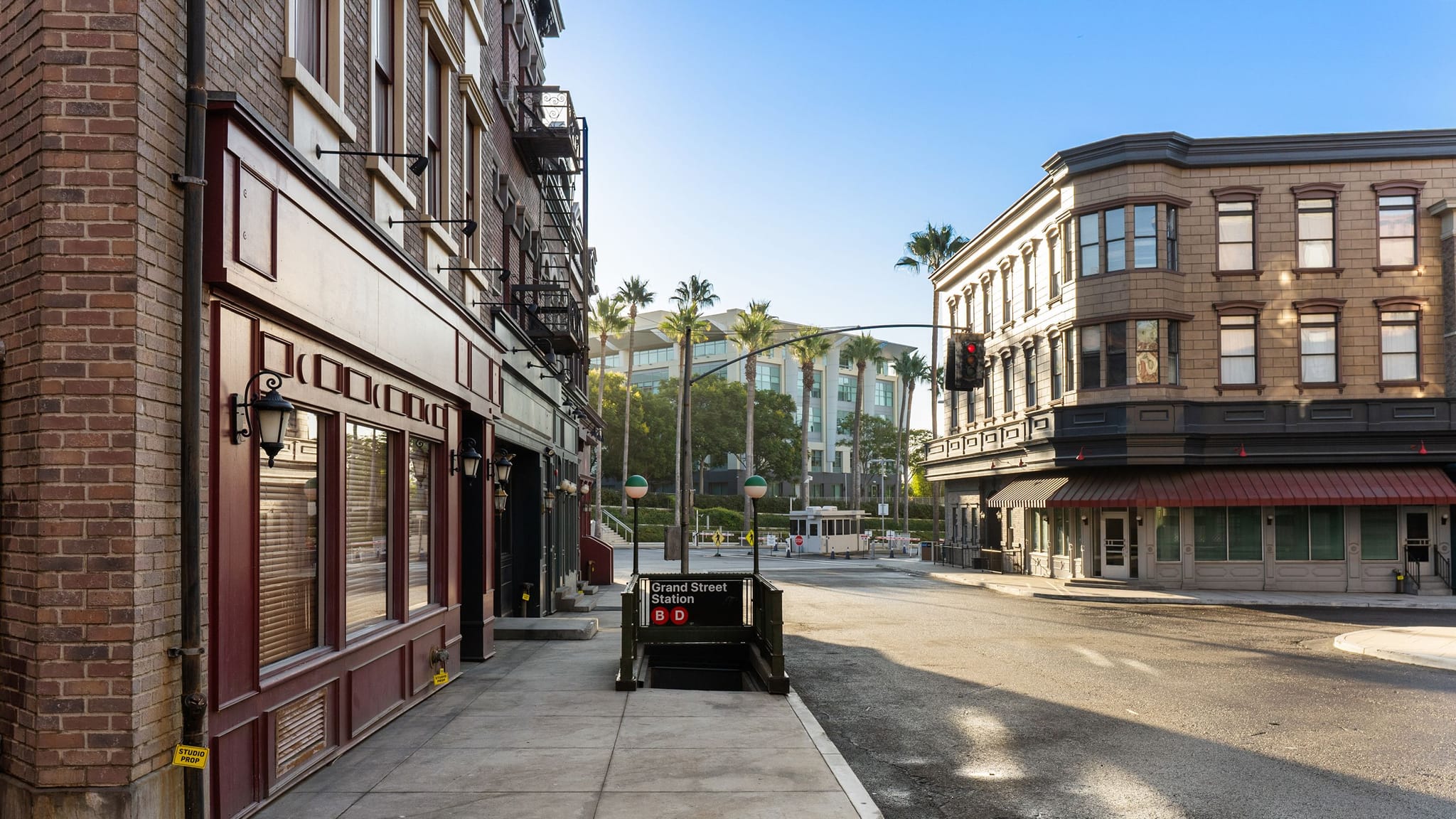 A street corner with brick buildings, a "Grand Street Station" subway entrance, and palm trees under a clear sky.