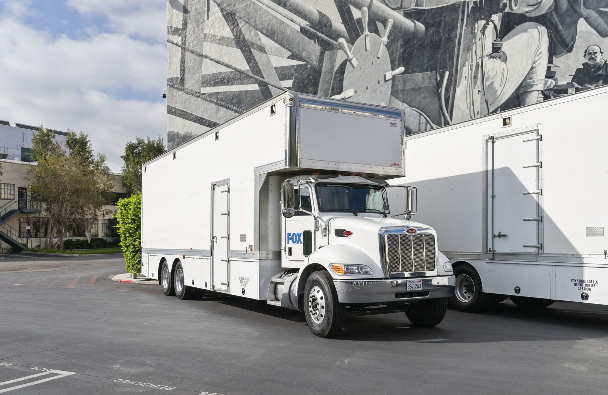 A white Fox broadcast truck with a trailer is parked in front of a building featuring a large black and white mural.