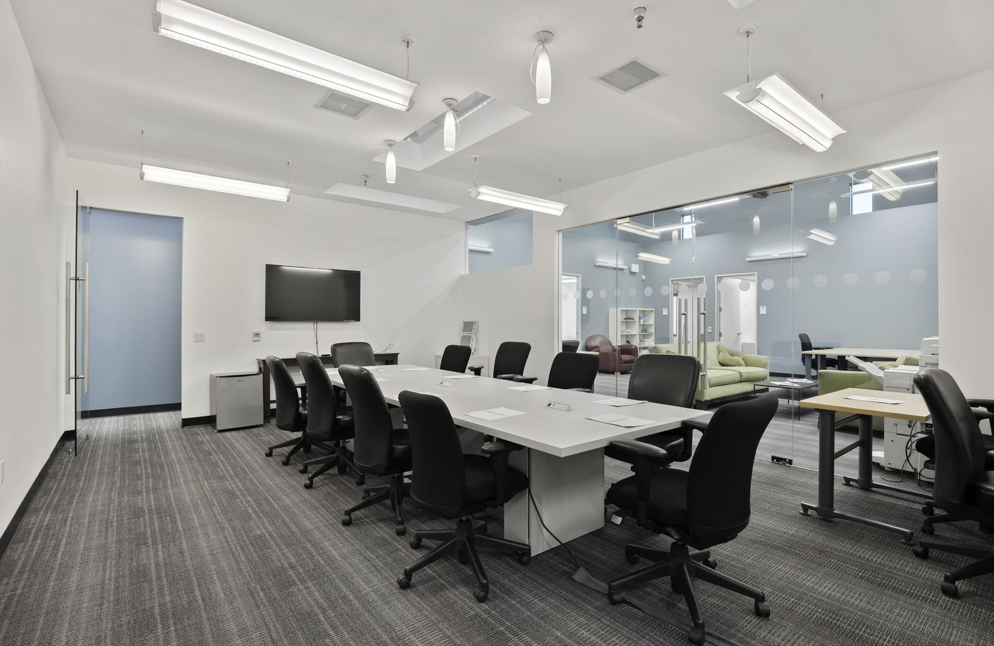 A modern conference room with a long white table, black chairs, a large TV, gray patterned carpet, and a glass wall looking into a lounge area.
