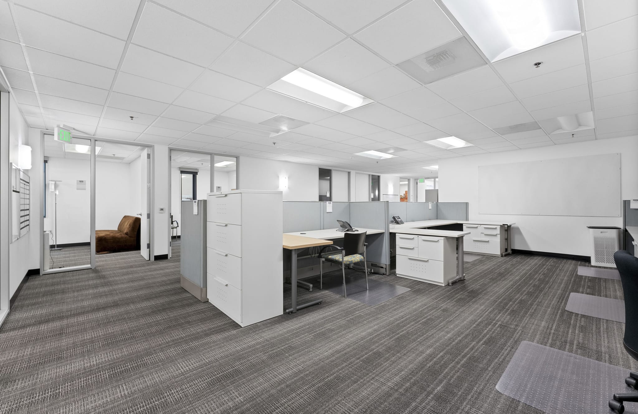 Empty open-plan office with multiple cubicles, patterned grey carpet, and glass-walled rooms.