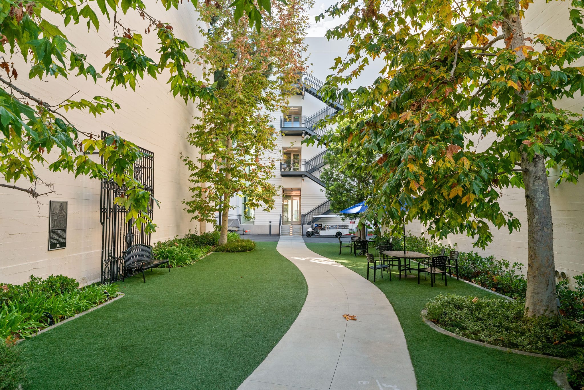 A winding concrete pathway through artificial green grass, flanked by trees and white building walls, with a bench and outdoor dining set.