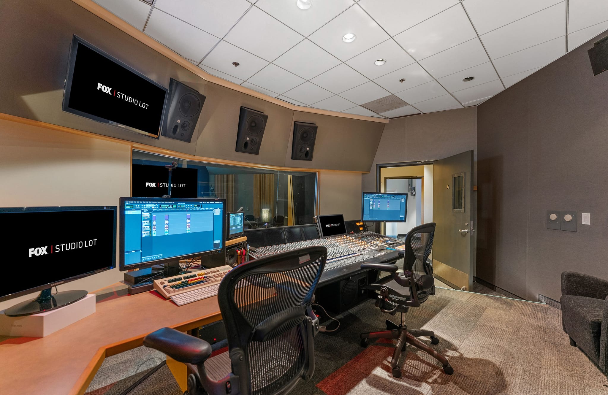 Control room of a modern audio studio with a mixing console, multiple monitors showing 'FOX | STUDIO LOT', and studio speakers.