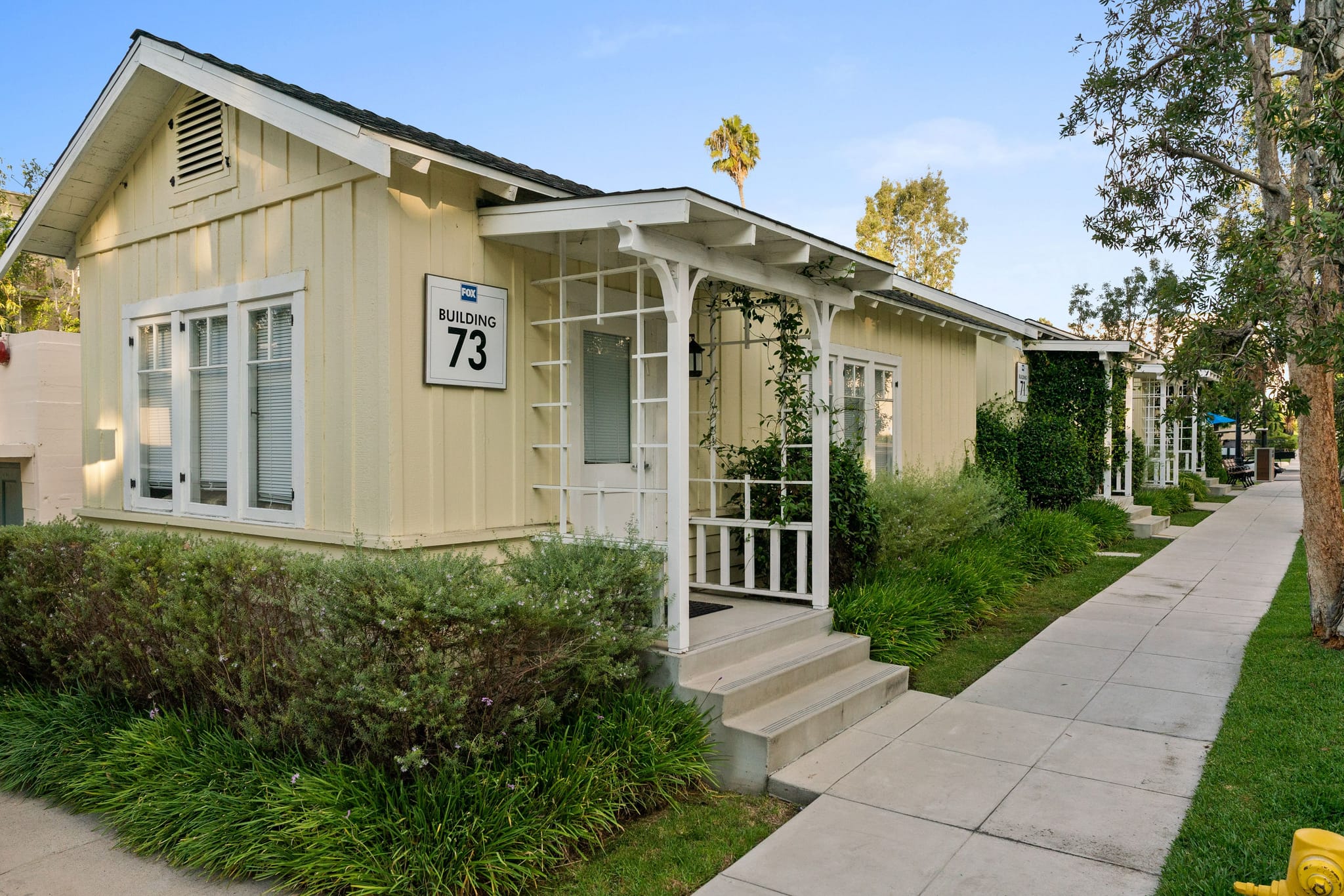 Pale yellow Building 73 with a white porch, part of a row of similar cottage-style buildings along a sidewalk with green landscaping.